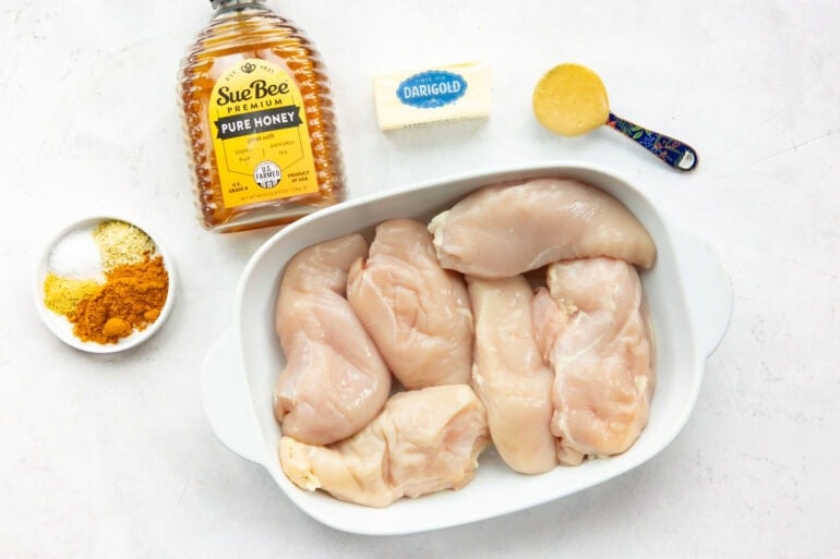 all the ingredients for honey curry chicken including raw chicken breasts in a baking dish with honey, curry powder, dijon mustard, butter and seasonings