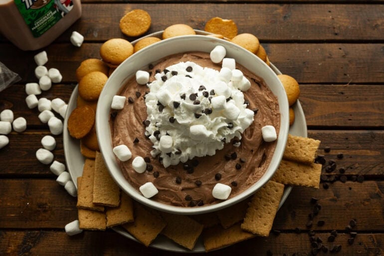 a large bowl of cream chocolate dip topped with mini marshmallows, mini chocolate chips surrounded by graham crackers and nilla wafers