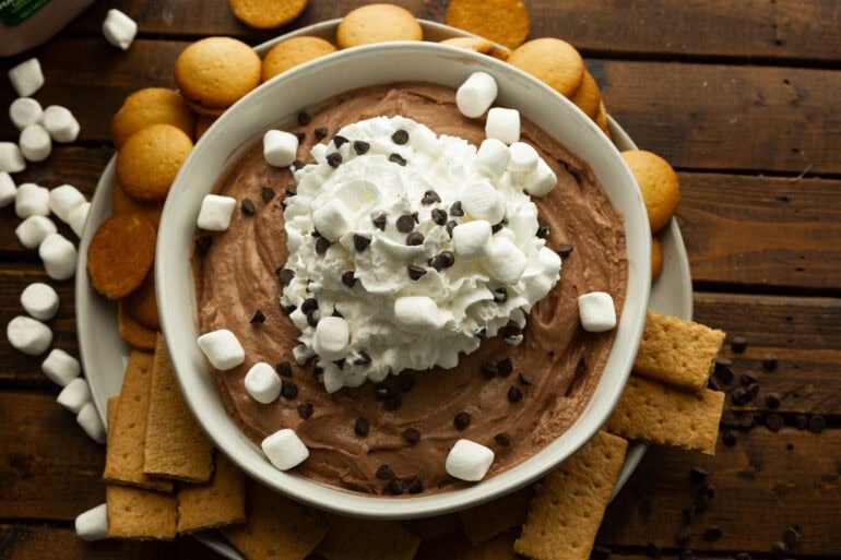 creamy hot cocoa dip in a large white bowl topped with whipped cream, mini marshmallows and mini chocolate chips
