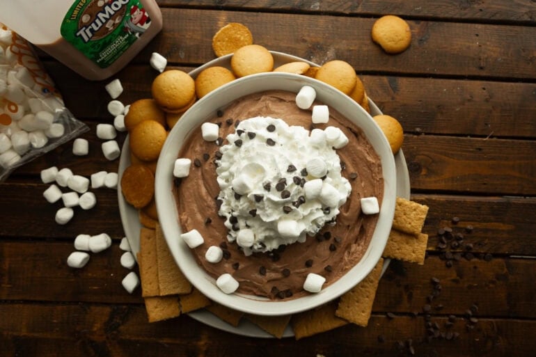 a large bowl of creamy chocolate dip topped with whipped cream, mini marshmallows and mini chocolate chips with nilla wafers and graham crackers