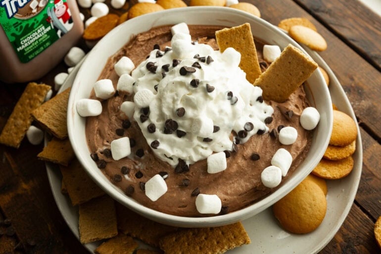 a large white bowl of hot cocoa dip tipped with whipped cream and sprinkled with mini chocolate chips and mini marshmallows with two graham crackers sticking out of it