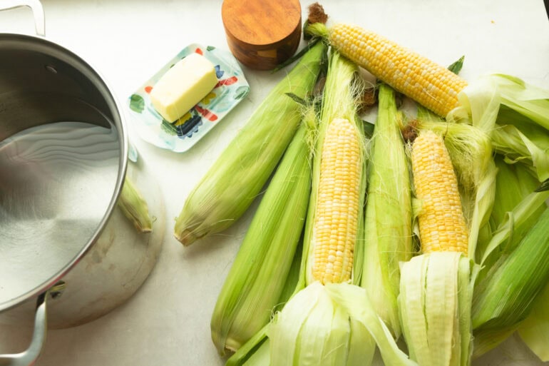 all the ingredients for the best corn on the cob including corn still in its husk, butter, salt and pepper and a large pot of water