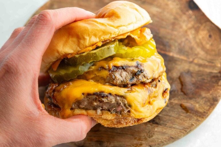 a hand holding a juicy double patty cheeseburger with dill pickle slices and a toasted bun