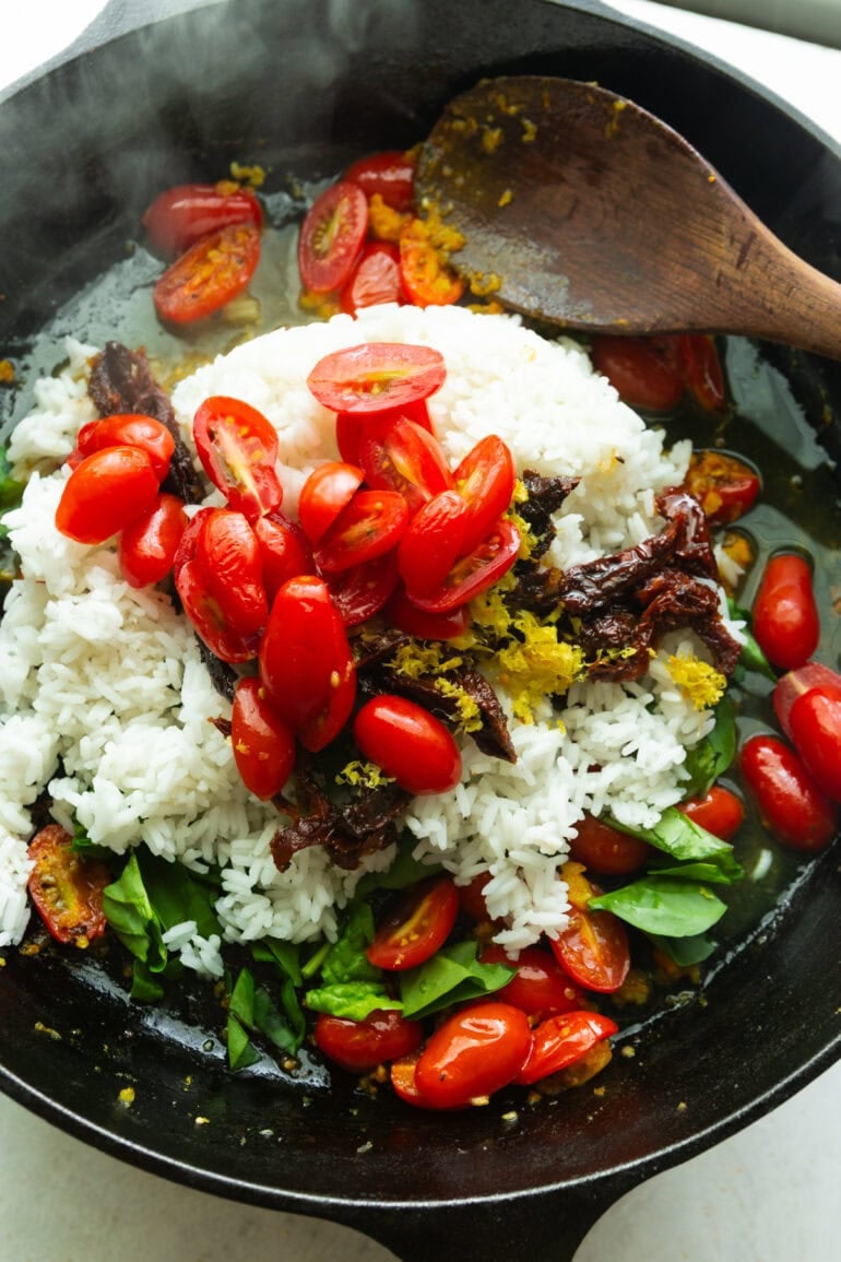 A cast Iron skillet is sizzling hot with rice, tomatoes, sun dried tomatoes, aromatic herbs, and olive oil/
