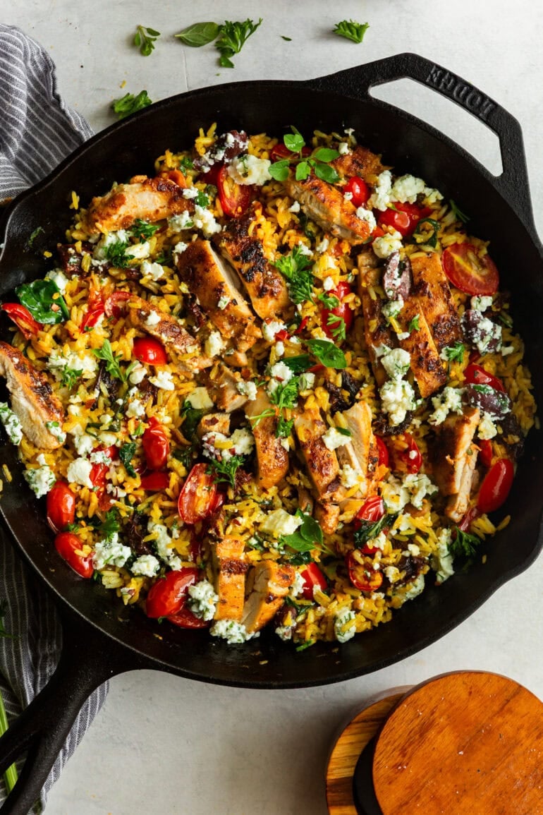 A cast iron pan with cooked Greek rice, tomatoes, herbs, feta, and grilled chicken