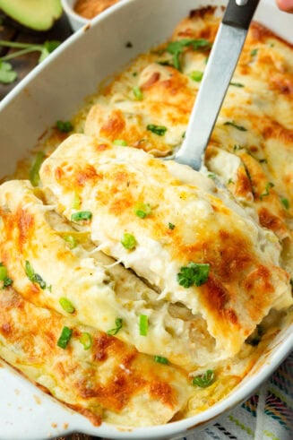 a photo of a cheesy chicken enchilada being lifted out of a pan full of golden cheesy enchiladas