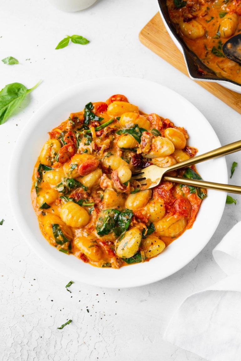 A photo of a white bowl full of creamy sun dried tomato gnocchi with a fork and spoon sitting in the bowl.