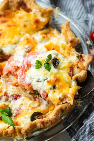 a photo of a slice of tomato pie being removed from a whole pie topped with melted cheese and fresh basil leaves