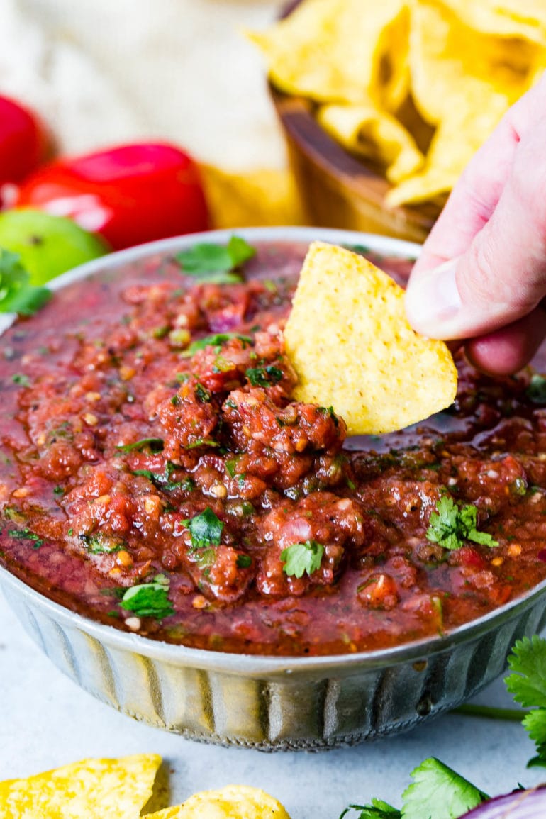A bowl of homemade salsa in which a tortilla chip is being dipped. The salsa is sprinkled with cilantro leaves and has cilantro and roma tomatoes and a lime in the background. There is also a wooden bowl of chips in the background.