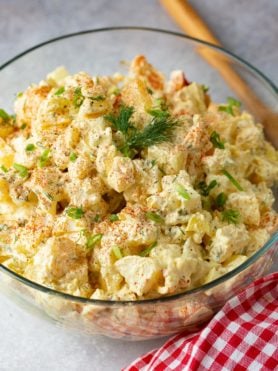 A glass serving bowl of potato salad. There are chunks of potato, chopped eggs, chopped pickles stirred together is a light dressing. The salad is topped with sprinkled paprika and fresh parsley. The bowl of salad is sitting next to a red checked napkin and a wooden spoon.