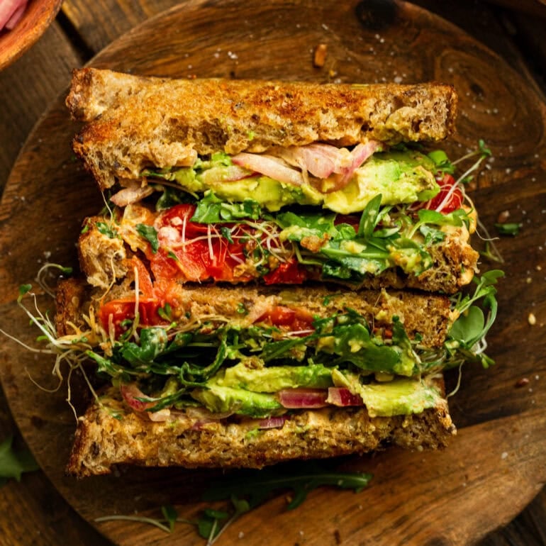 a photo of an avocado sandwich with toasted multigrain bread, bean sprouts, red onions, avocado, arugula, etc.