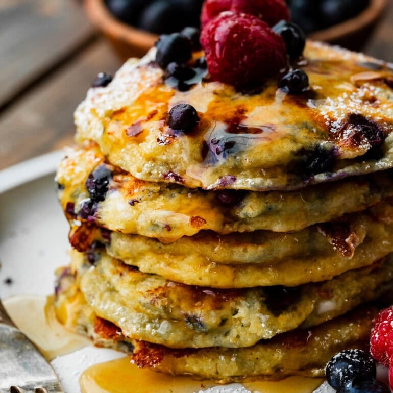 a photo of a stack of blueberry cottage cheese pancakes covered in maple syrup and topped with fresh raspberries and blueberries