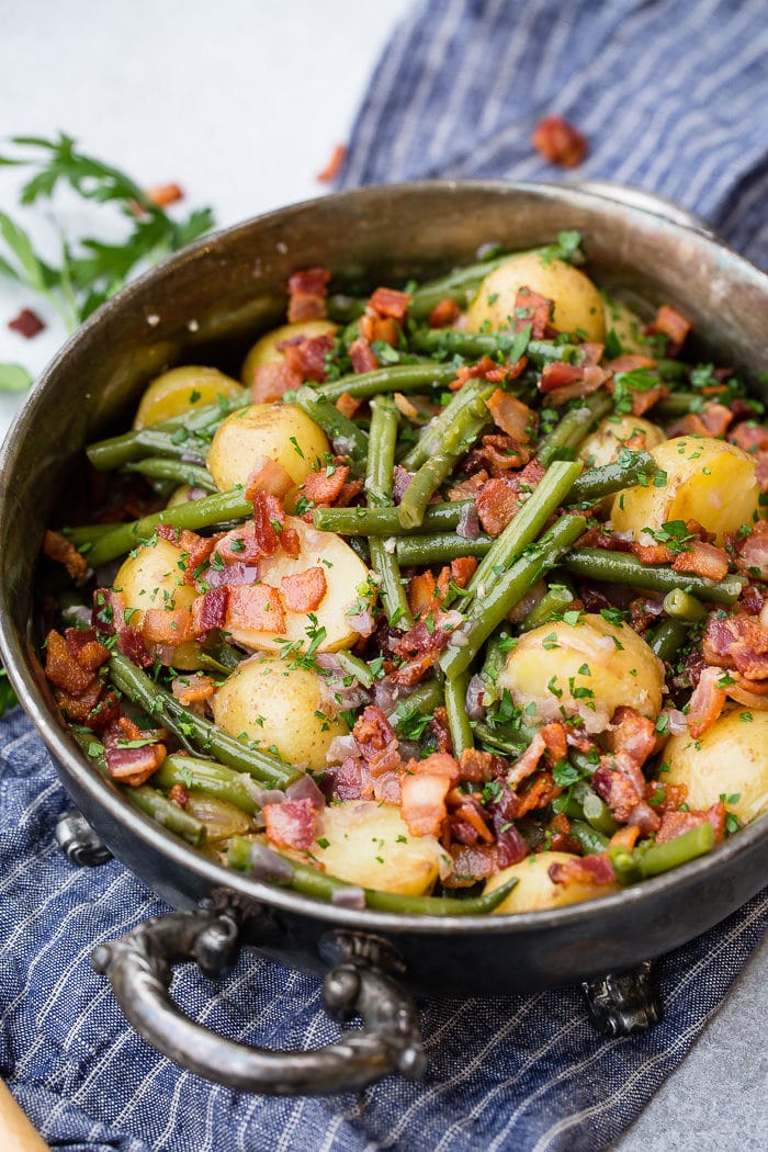 A photo of a silver serving dish sitting on a blue cloth napkin that is full of cooked fresh green beans, potatoes, and bacon pieces sprinkled with fresh parsley.