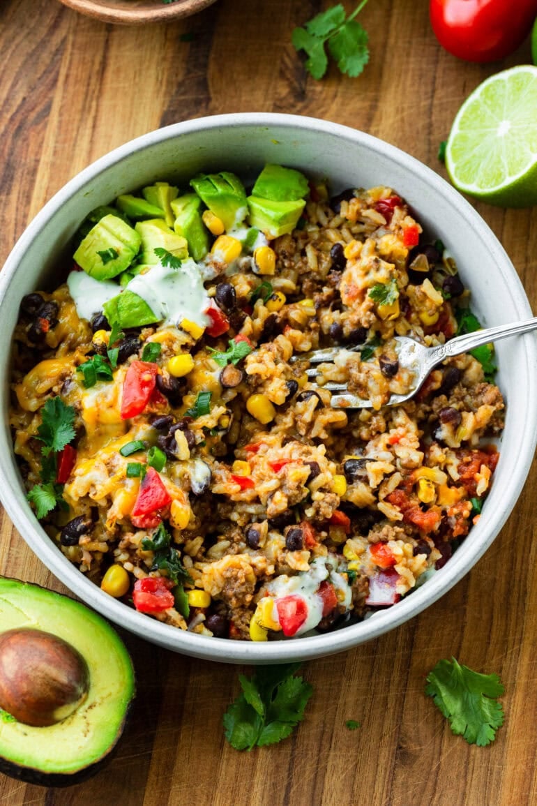 a bowl full of ground beef and rice mixed with black beans, diced tomatoes, melted cheese, avocado and sour cream