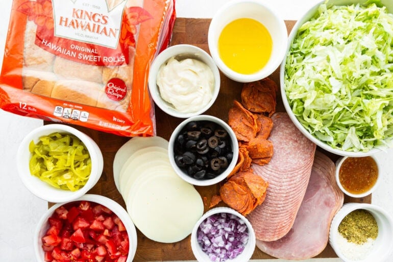 a photo of all the ingredients for italian sliders including Kings Hawaiian rolls, slices of provolone, sliced meats, chopped lettuce tomatoes, red onions, olives, etc.