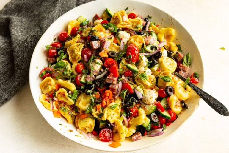 a large white bowl of tortellini pasta salad including black olives, diced red onions, halved tomatoes, cucumbers, salami, all mixed with Italian dressing