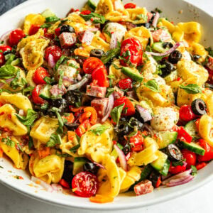 a large white bowl of tortellini pasta salad with black olives, red onions, cherry tomatoes, mozzarella balls and herbs