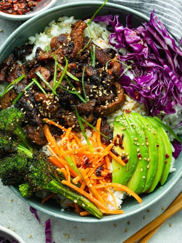 a rice bowl topped with roasted broccoli, red cabbage, shredded carrots, sliced avocado and strips of korean beef