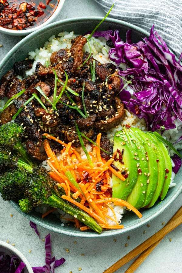 a rice bowl topped with roasted broccoli, red cabbage, shredded carrots, sliced avocado and strips of korean beef