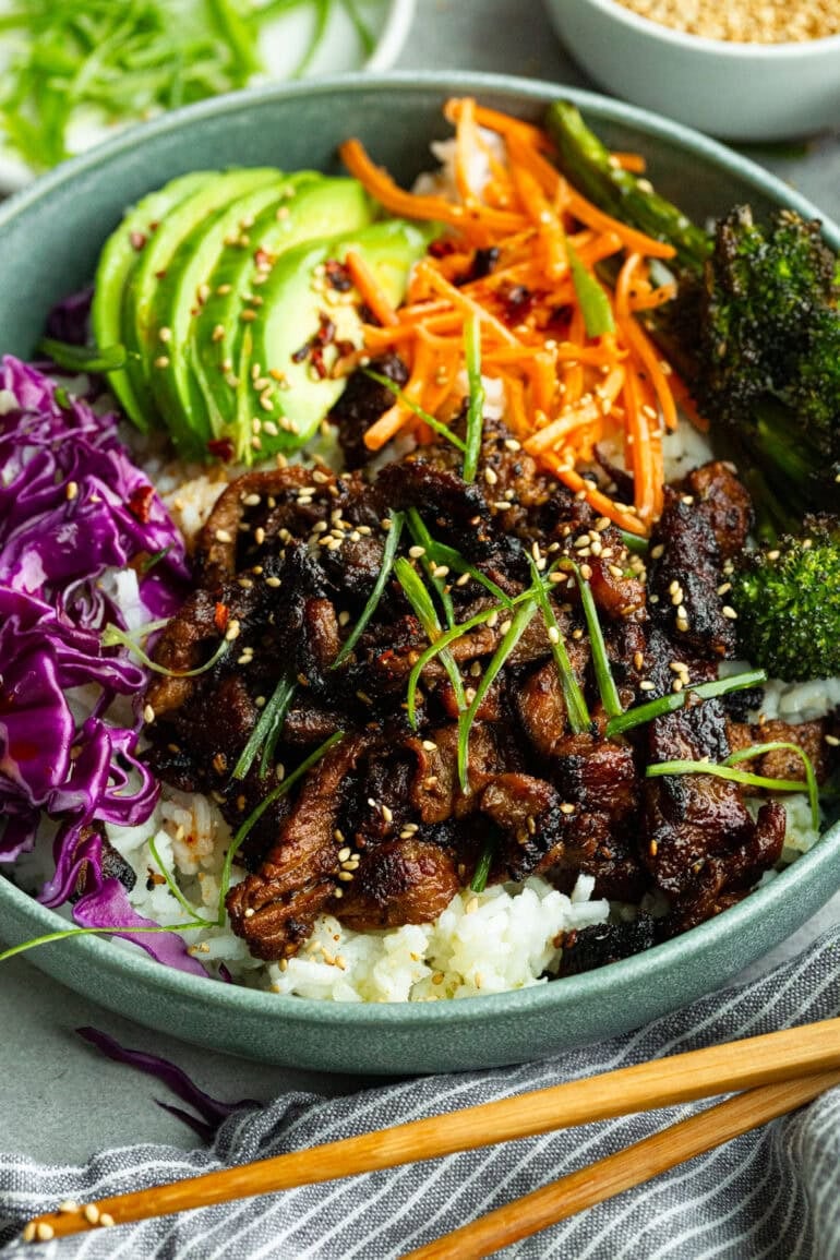 a korean beef rice bowl topped with sesame seeds, matchstick carrots, shredded purple cabbage, and sliced avocados