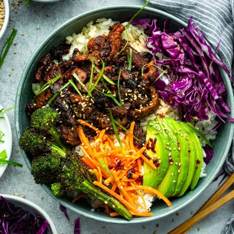 a photo of a bowl of fluffy white rice topped with saucy strips of beef, roasted broccoli, shredded purple cabbage, matchstick carrots and sliced avocado