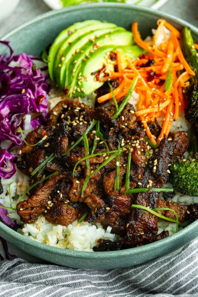 a bowl of white rice topped with strips of sauteed beef, roasted broccoli, shredded carrots and purple cabbage with sliced avocados on the side
