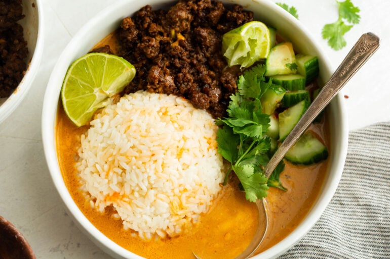 a mound of rice surrounded by a rich coconut gochujang broth and ground beef, cucumbers, cilantro and lime wedge