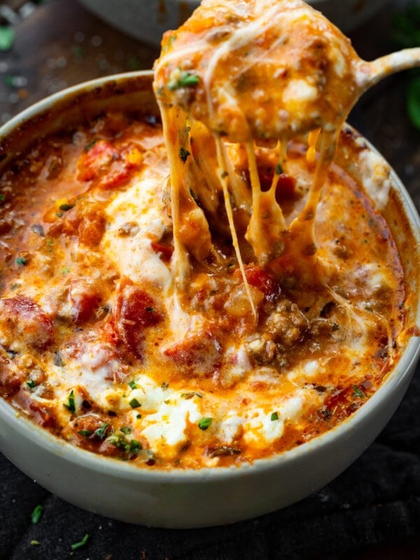 a photo of a bowl of lasagna soup topped with melted cheese and someone pulling a spoon out of the bowl with cheese stringing from it.