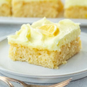 a photo of a square slice of moist lemon cake topped with lemon cream cheese frosting and a thin slice of fresh lemon on top