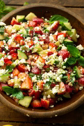 a photo of a large wooden bowl full of strawberry cucumber salad with feta and fresh herbs