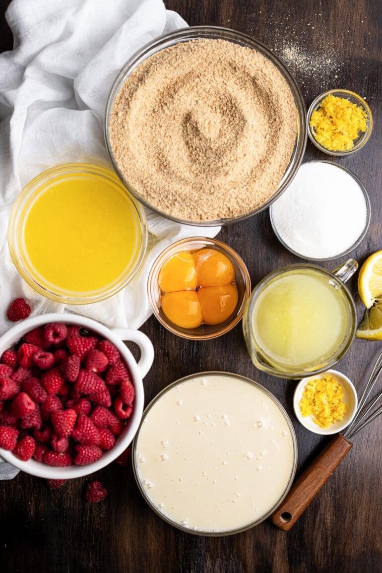 all the ingredients for lemon raspberry bars in separate small bowls including whole raspberries, butter, egg yolks, sweetened condensed milk, lemon juice and zest, sugar, and 
graham cracker crumbs