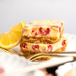 a photo of two creamy lemon raspberry bars with graham cracker crust stacked on each other on a small dessert plate with a lemon wedge next to them
