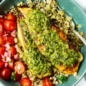 a photo of two golden tilapia filets topped with bright pesto sauce on a bed of quinoa with grape tomatoes on the side