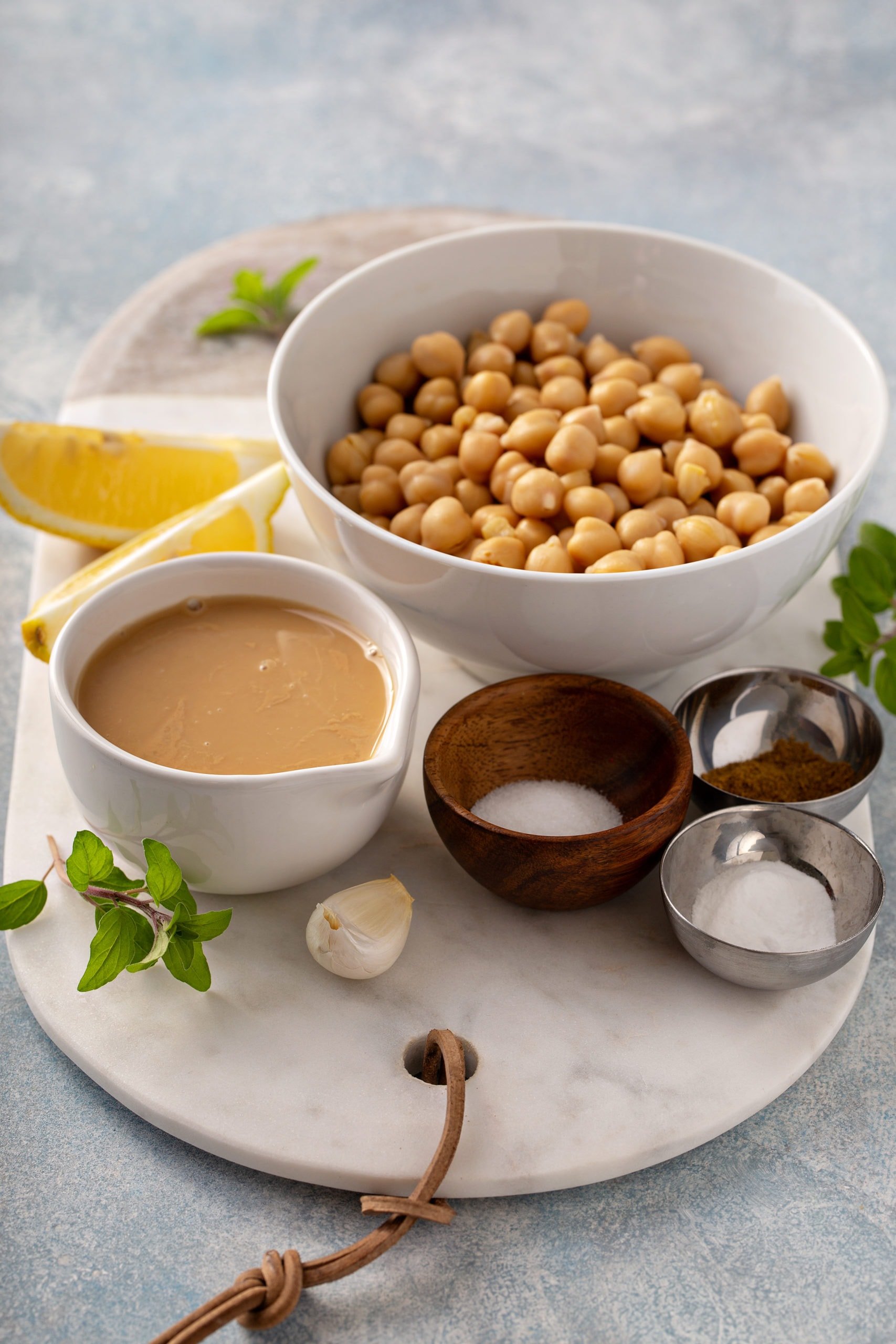 A large bowl of chick peas, smaller bowls of sea salt, cumin and baking soda with a cup of Tahini and a clove of garlic and lemon wedges.