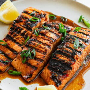 three grilled salmon filets sitting on a white plate sitting in a saucy marinade
