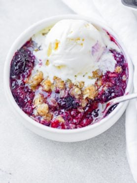 a photo of a single serving of marionberry crisp in a white bowl with a silver spoon in it topped with a scoop of vanilla ice cream that is starting to melt into the crisp.