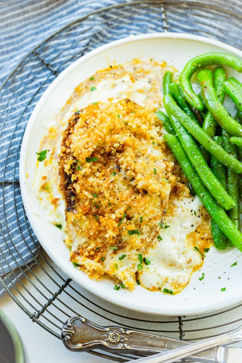 a photo of a single panko covered chicken breast sitting in a creamy sauce with a serving of fresh green beans on the side.