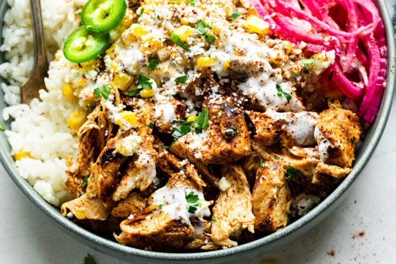 a street corn chicken rice bowl with seasoning chicken breasts pieces, creamy elote, rice, sliced jalapenos, and pickled red onions