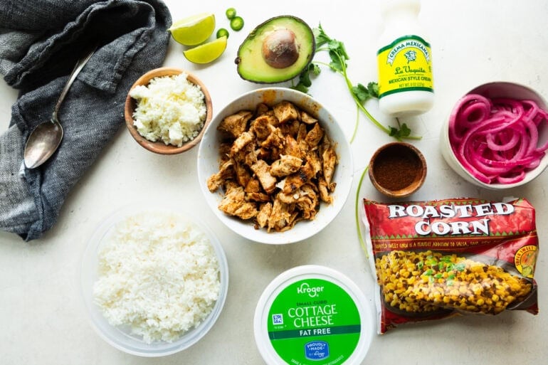all the ingredients for street corn chicken rice bowls including seasoned chicken chunks, cottage cheese, roasted corn, avocado, rice, mexican crema, and seasonings