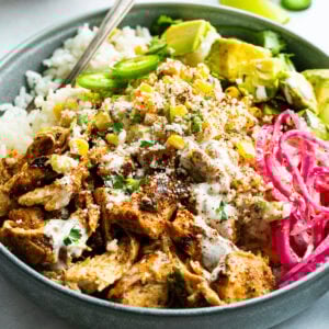 a mexican chicken rice bowl topped with creamy elote, picked red onions, avocado and jalapeno slices