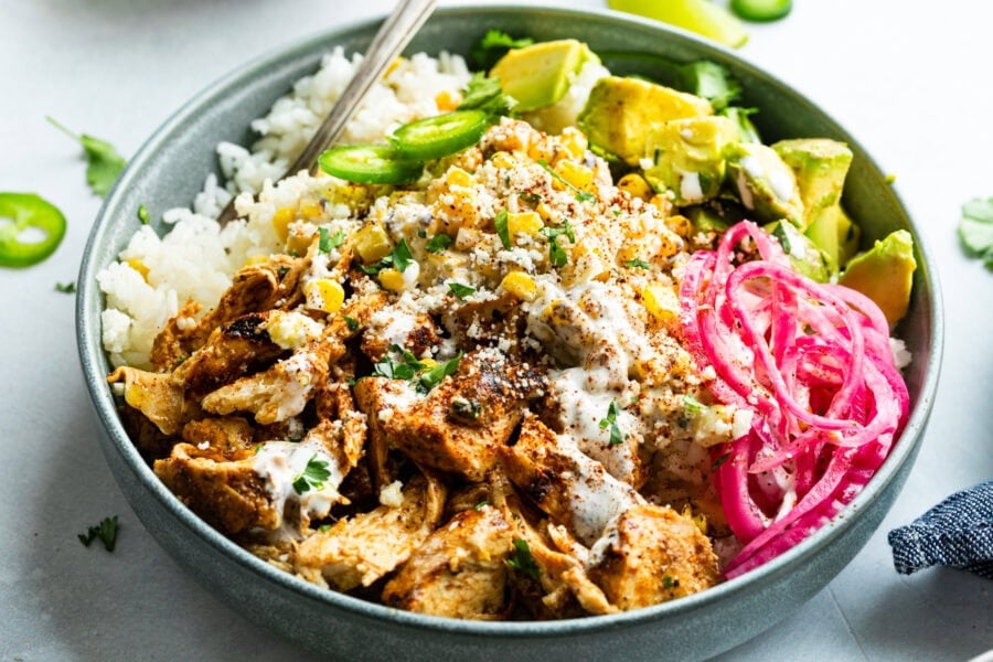 a mexican chicken rice bowl topped with creamy elote, picked red onions, avocado and jalapeno slices
