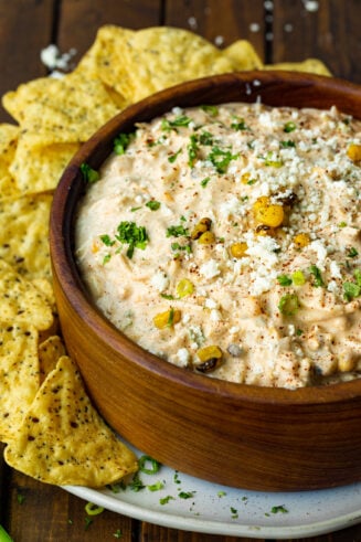 a photo of a large wooden bowl full of Mexican corn dip with tortilla chips scattered around the bowl.