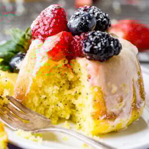 A photo of a mini lemon poppy seed bundt cake on on a silver wire cooling rack with a lemon glaze on top and fresh berries.