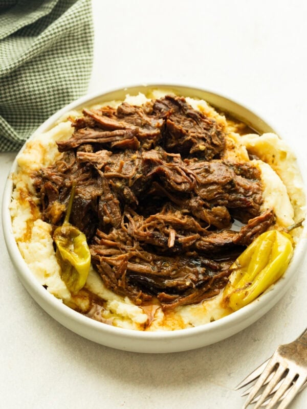 a large serving bowl full of juicy, tender beef served over the top of creamy mashed potatoes and whole pepperoncicnis around the bowl