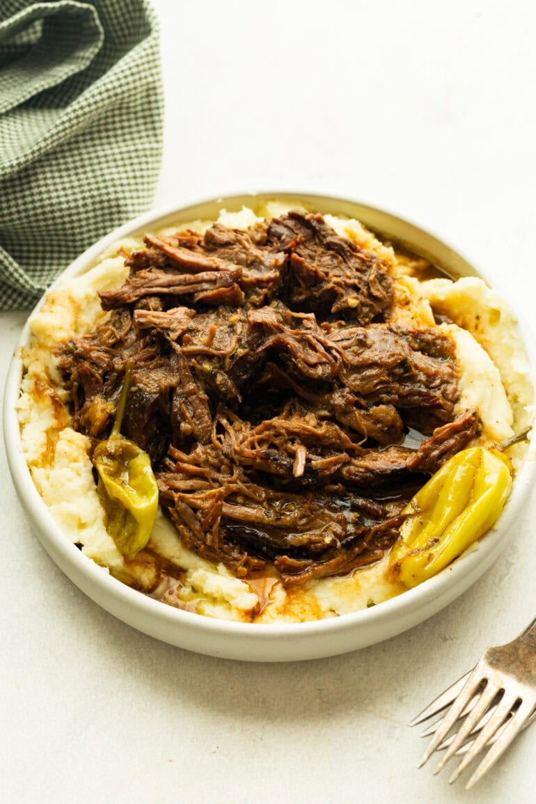 a large serving bowl full of juicy, tender beef served over the top of creamy mashed potatoes and whole pepperoncicnis around the bowl