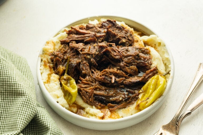 a large serving bowl full of creamy mashed potatoes topped with juicy, tender shredded beef with pepperoncinis scattered around it