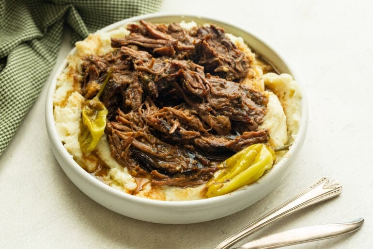 a large serving bowl of creamy mashed potatoes with juicy shredded pot roast on top and a few whole pepperoncinis