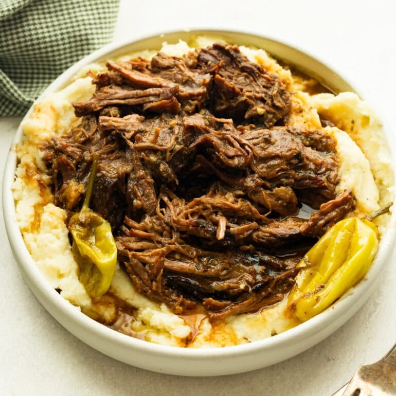 a large white serving bowl full of creamy mashed potatoes topped with juicy shredded pot roast with whole pepperoncinis scattered around the bowl
