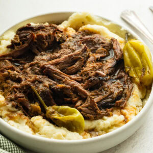 a large white serving bowl full of creamy mashed potatoes topped with juicy shredded pot roast with whole pepperoncinis scattered around the bowl