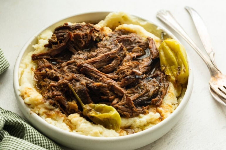 a large white serving bowl full of creamy mashed potatoes topped with juicy shredded pot roast with whole pepperoncinis scattered around the bowl